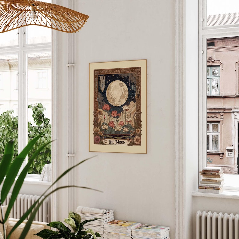 May include: A framed art print depicting a full moon, lotus flowers, and celestial imagery. The artwork has a vintage style with a beige border and the words "THE MOON" at the bottom. The print is displayed on a white wall near a window.