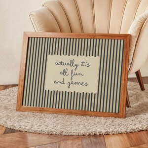 May include: A framed print with a wooden frame. The print has a striped background in dark green and beige. A beige, scalloped-edge rectangle in the center displays the text "actually it's all fun and games" in black script.