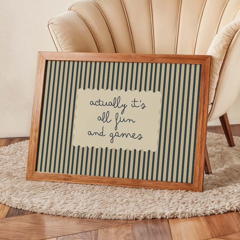 May include: A framed print with a wooden frame. The print has a striped background in dark green and beige. A beige, scalloped-edge rectangle in the center displays the text "actually it's all fun and games" in black script.