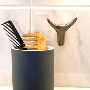 May include: A gray ceramic bathroom cup with a black comb and a wooden comb inside. A silver bull head hook is mounted on the wall behind the cup.