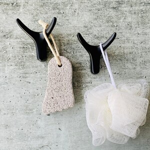 May include: Two black bull-shaped hooks mounted on a wall. A pumice stone is hanging from one hook and a white bath pouf is hanging from the other.