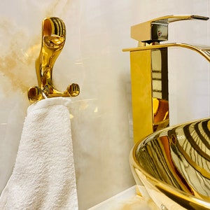 Könnte beinhalten: Ein vergoldetes Badezimmerwaschbecken mit passendem Wasserhahn und Handtuchhaken. Das Waschbecken ist oval geformt und hat eine glatte, reflektierende Oberfläche.