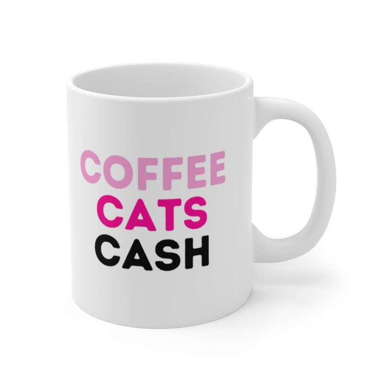 May include: White ceramic coffee mug with the text "COFFEE CATS CASH" in pink and black lettering.