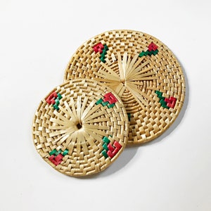 May include: Two woven straw coasters with red and green floral designs. The coasters are round and have a natural, rustic look.