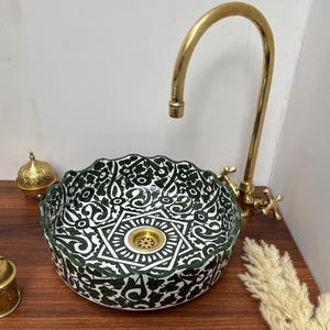 Puede incluir: Un lavabo de cerámica blanca con un patrón geométrico verde y blanco. El lavabo tiene una forma redonda con un borde festoneado. Un grifo dorado está montado sobre el lavabo.