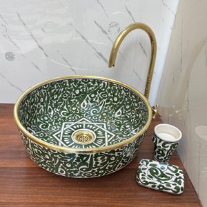 May include: A green and white ceramic bathroom sink with a gold rim and matching soap dish and cup. The sink has an intricate floral pattern and a gold faucet.