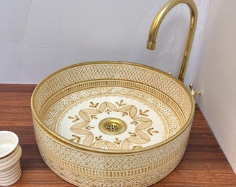 Puede incluir: Un lavabo de cerámica blanca con un patrón geométrico dorado y un diseño de hojas doradas. El lavabo es redondo y tiene un grifo de latón.