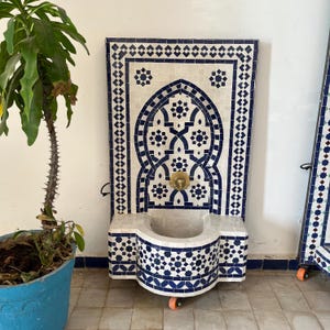 Może przedstawiać: Biała ceramiczna fontanna ścienna z niebieskimi i białymi wzorami z mozaiki. Fontanna ma dyszę i misę. Fontanna jest na kółkach i znajduje się przed białą ścianą.