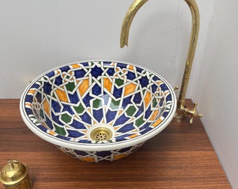 May include: A decorative ceramic sink with a geometric pattern in blue, orange, and green. The sink is paired with a gold-colored faucet. A small, gold-colored container sits on the wooden surface next to the sink.