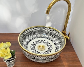 Può includere: Un lavandino in ceramica bianca con un motivo geometrico nero e oro. Il lavandino è rotondo e ha un bordo dorato. Un rubinetto dorato è montato sopra il lavandino.