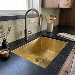 Può includere: Un lavello da cucina in ottone spazzolato con rubinetto a canna alta abbinato, incassato in un piano di lavoro grigio scuro. Accanto si trovano una piccola ciotola in legno con sale, un vaso in ceramica scura con lavanda e un panno in lino.