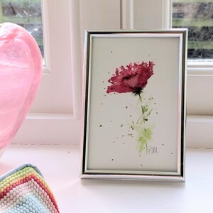 May include: A framed watercolour painting of a red poppy flower with green accents. The artwork is in a silver frame and features a delicate, artistic style. The painting is set against a white background.