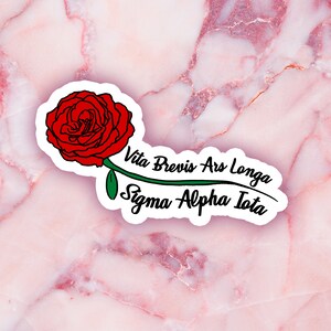 May include: A sticker featuring a red rose with a green stem and leaf. The text "Vita Brevis Ars Longa Sigma Alpha Iota" is written in black script. The sticker is on a pink marble background.