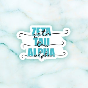 May include: A white sticker with the words "ZETA TAU ALPHA" in a teal color. The word "ZETA" is stacked above "TAU" and "ALPHA". The words are in a bold, sans-serif font. Black, swirly lines are above and below the words.