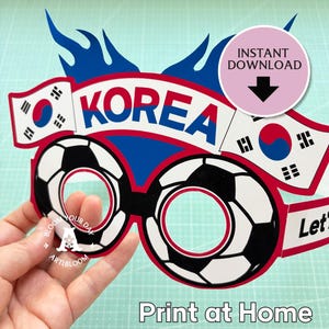 May include: A paper party glasses cutout featuring two soccer ball frames, the text KOREA, two South Korean flags, and blue flame accents. The design includes a pink circular badge with the text INSTANT DOWNLOAD and a downward arrow.