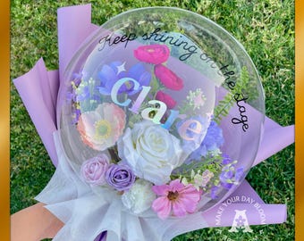Peut inclure: Un ballon bulle transparent rempli de fleurs artificielles colorées, dont des roses blanches, des fleurs roses et violettes et de la verdure. Le ballon porte le nom "Clare" et la phrase "Keep shining on the stage". Le bouquet est enveloppé de papier violet et blanc.