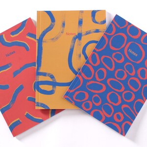 May include: Three notebooks with different abstract designs. The notebooks are all the same size and have a red, blue, and yellow colour scheme. The word "Notes" is printed on each notebook.