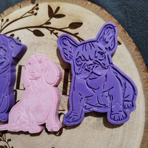 Op de afbeelding: Drie hondenkoekjessnijders in paars en roze. De hondenrassen zijn een Franse Bulldog, een Teckel en een ander ras. De uitstekers liggen op een houten schijf met een decoratief bladerenpatroon.