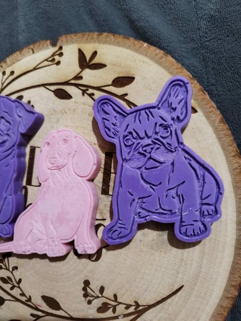 Op de afbeelding: Drie hondenkoekjessnijders in paars en roze. De hondenrassen zijn een Franse Bulldog, een Teckel en een ander ras. De uitstekers liggen op een houten schijf met een decoratief bladerenpatroon.
