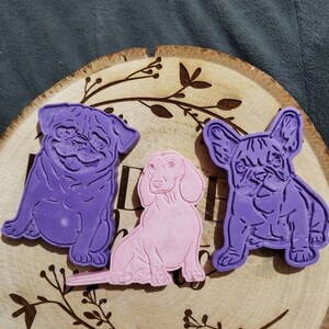 Op de afbeelding: Drie hondenmallen liggen op een houten schijf met een bladerenpatroon. De mallen tonen een paarse mopshond, een roze teckel en een paarse Franse bulldog. De houten schijf heeft een natuurlijke houtnerf.