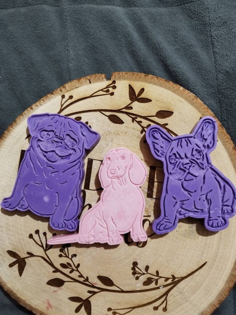 Op de afbeelding: Drie hondenmallen liggen op een houten schijf met een bladerenpatroon. De mallen tonen een paarse mopshond, een roze teckel en een paarse Franse bulldog. De houten schijf heeft een natuurlijke houtnerf.