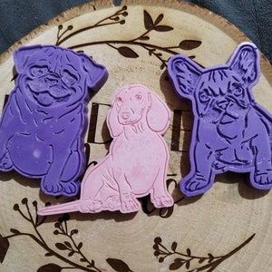 Op de afbeelding: Drie hondenmallen in paars en roze zijn gerangschikt op een houten schijf met een decoratief bloemmotief. De mallen omvatten een mopshond, een teckel en een Franse bulldog.