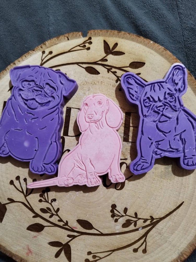 Op de afbeelding: Drie hondenmallen in paars en roze zijn gerangschikt op een houten schijf met een decoratief bloemmotief. De mallen omvatten een mopshond, een teckel en een Franse bulldog.