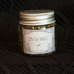 May include: A small glass jar with a silver lid, filled with a green and brown mixture. A white label with the words "D.u.Me oil" is affixed to the jar. The jar is set against a dark, textured background.