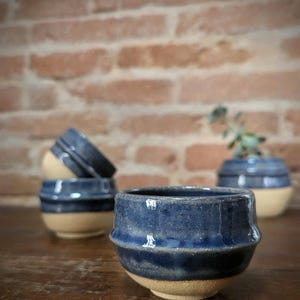 May include: A collection of handmade ceramic bowls. The bowls are a deep blue color on the top half and a tan color on the bottom half. The bowls are sitting on a wooden surface.