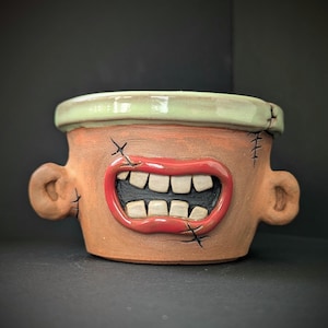 Może przedstawiać: Ręcznie robiona ceramiczna donica o unikalnym, potwornym designie. Donica posiada szerokie usta z czerwonymi wargami i białymi zębami, detale w postaci szwów oraz małe uchwyty w kształcie uszu. Obręcz jest w kolorze jasnozielonym.