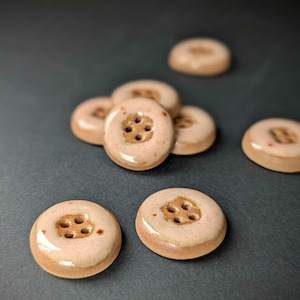 May include: A collection of handmade ceramic buttons in a soft, muted peach color. Each button is round with four holes and a slightly raised center. The buttons have a glossy finish and are scattered on a dark gray surface.
