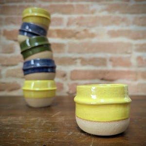 May include: A stack of handmade ceramic bowls in various colors, including yellow, blue, and green. The bowls have a textured, light-colored base and a smooth, glossy upper section. One yellow bowl sits in front of the stack on a wooden surface.