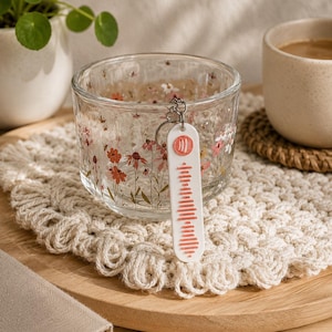Könnte beinhalten: Ein durchsichtiger Glas-Kerzenhalter mit floralem Design und einem weißen Schlüsselanhänger mit Musikcode. Der Kerzenhalter steht auf einem cremefarbenen gehäkelten Untersetzer, mit einer Kaffeetasse und einer kleinen Pflanze im Hintergrund.