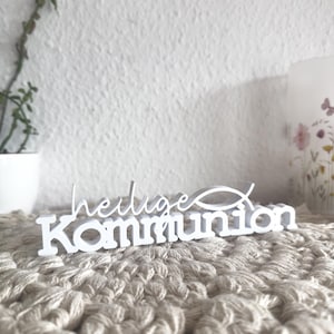 Puede incluir: Un letrero de madera blanco con las palabras alemanas "Heilige Kommunion" y un símbolo de pez. El letrero está colocado sobre una superficie beige texturizada, con una pared blanca y una planta en maceta en el fondo.
