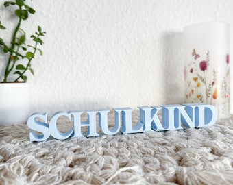 Könnte beinhalten: Hellblaue Holzbuchstaben, die das Wort "SCHULKIND" auf einer hellbraunen, strukturierten Oberfläche buchstabieren.