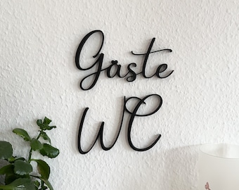 Könnte beinhalten: Schwarzes Holzschild mit den Worten "Gäste WC" in Kursivschrift. Das Schild ist an einer weißen Wand befestigt.