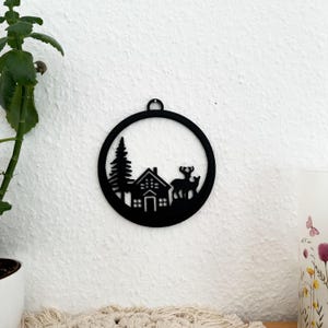 May include: Black metal ornament featuring a silhouette of a cabin, a pine tree, and two deer in a circular frame.