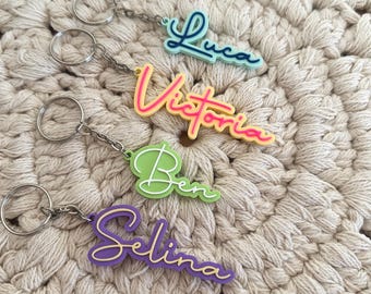 Puede incluir: Cuatro llaveros personalizados con nombres en letra cursiva. El superior es azul claro con texto azul oscuro que dice Luca. El segundo es amarillo con texto rosa que dice Victoria. El tercero es verde claro con texto blanco que dice Ben. El inferior es morado con texto amarillo que dice Selina.