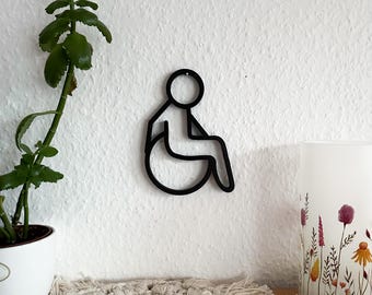 Könnte beinhalten: Schwarze Metallumrisszeichnung einer Person im Rollstuhl. Das Symbol ist an einer weißen Wand montiert.