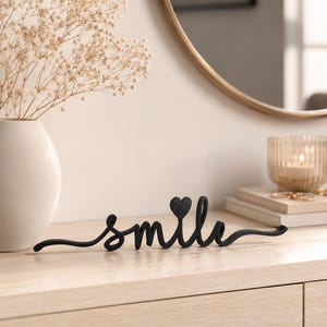 Könnte beinhalten: Schwarze Metallskulptur des Wortes "smile" mit einem herzförmigen Akzent über dem Buchstaben "i". Die Skulptur steht auf einer hellen Holzoberfläche, im Hintergrund eine Vase mit Trockenblumen und eine Kerze.