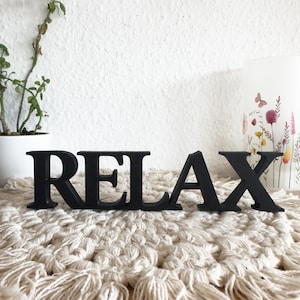 Puede incluir: Letras de madera negras que deletrean la palabra "RELAX". Las letras están colocadas sobre una superficie de macramé color crema. Una vela blanca con diseños florales y una planta en maceta están en el fondo.