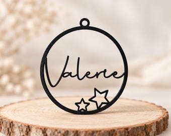 Puede incluir: Un adorno circular negro que presenta el nombre Valerie en una fuente cursiva. El diseño incluye dos estrellas de contorno abierto en la parte inferior y un pequeño lazo en la parte superior para colgar. El objeto es de material plano y sólido.