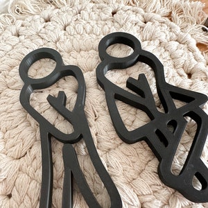 Puede incluir: Dos figuras de personas de madera recortada en negro, de pie una al lado de la otra. Las figuras son estilizadas y tienen un dise&ntilde;o simple. Est&aacute;n colocadas sobre una superficie de crochet blanco.