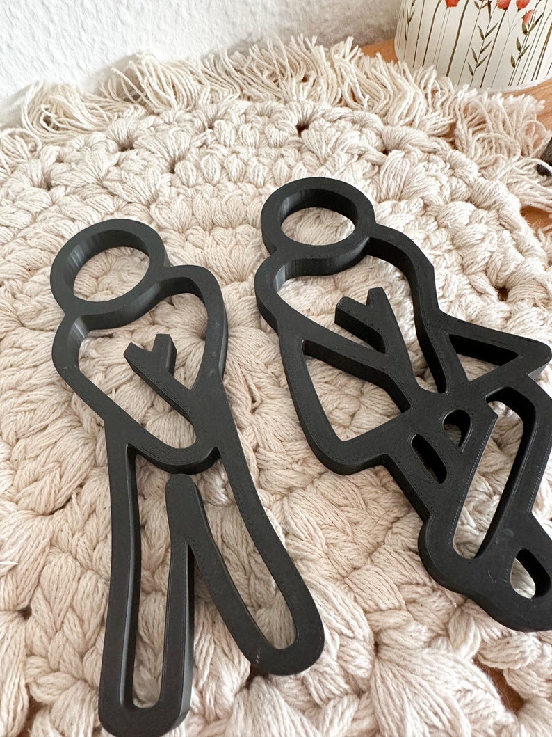 Puede incluir: Dos figuras de personas de madera recortada en negro, de pie una al lado de la otra. Las figuras son estilizadas y tienen un dise&ntilde;o simple. Est&aacute;n colocadas sobre una superficie de crochet blanco.