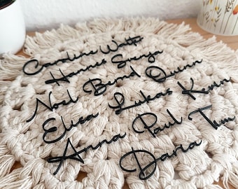 May include: Black laser-cut name place cards in a cursive font, arranged on a cream-colored macrame placemat. Names include Caroline, Hannes, Svenja, Bernd, Quinn, Noel, Lukas, Kevin, Elsa, Anna, Paul, Tim, and Daria. A decorative plant and vase are in the background.