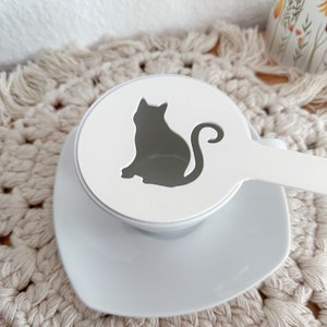 Könnte beinhalten: Weiße Keramik-Kaffeenschablone mit einer Silhouette einer Katze. Die Schablone hat einen Griff für einfache Verwendung.