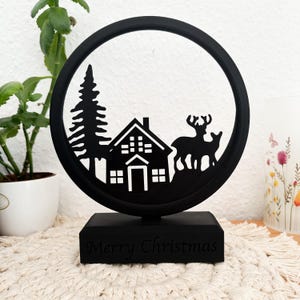 May include: Black silhouette Christmas ornament featuring a house, a pine tree, and two deer. The ornament is on a black base with the words "Merry Christmas" inscribed on the front.