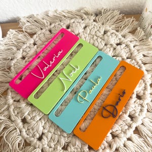 Könnte beinhalten: Vier rechteckige Namensschilder aus Acryl in den Farben Pink, Limettengrün, Hellblau und Orange. Jedes Schild hat einen horizontalen Schlitz und einen erhabenen, in Schreibschrift gestalteten Namen: Valeria, Noel, Paula und Dario.