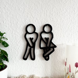 Puede incluir: Decoraci&oacute;n de pared de silueta negra de un hombre y una mujer en un estilo de ba&ntilde;o. Las siluetas est&aacute;n recortadas de un material negro y montadas en una pared blanca.
