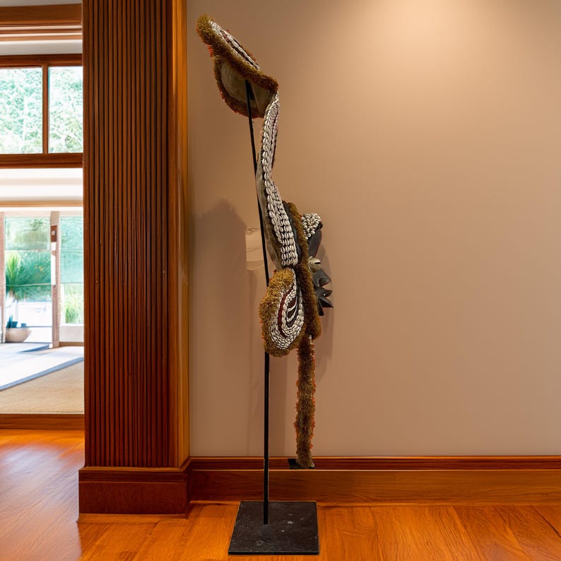 May include: A large, intricately carved wooden mask with white and brown accents. The mask is mounted on a black metal stand and has a long, flowing tail made of brown feathers.
